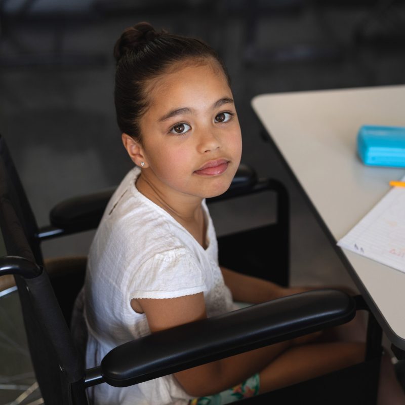 High angle view of disabled schoolgirl looking at camera and sitting at desk in classroom of elementary school