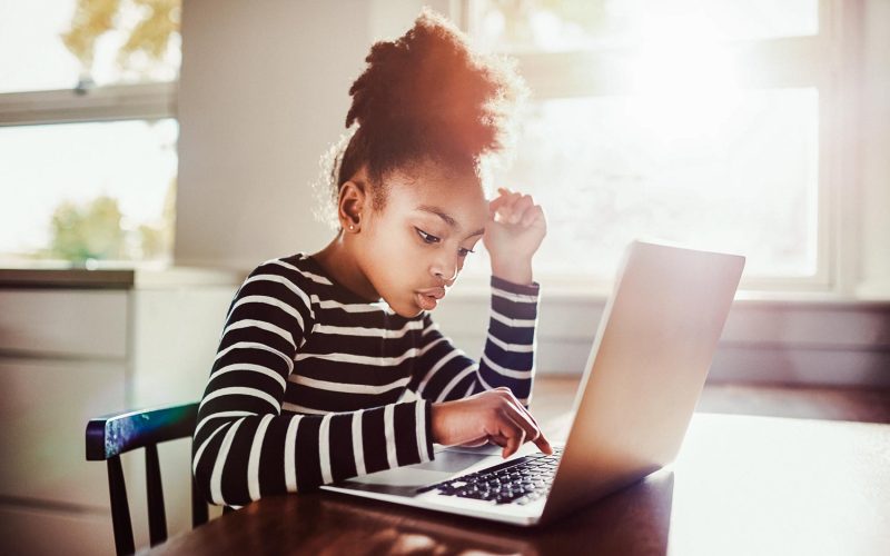 Young girl sitting at the dining table at home working on her homework from school typing out an answer on a laptop computer, e-learning concept