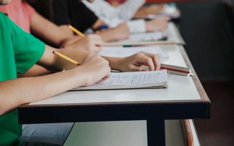Midsection of high school students writing on paper at desk in classroom