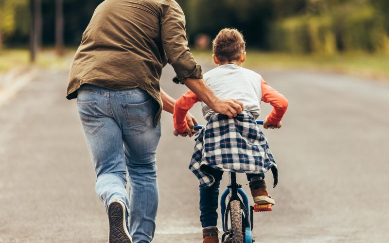 Rear view of a boy riding a bicycle while his father runs along holding the kid. Father teaching his son to ride a bicycle.