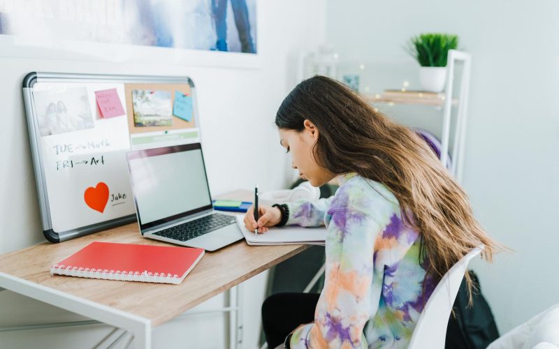 Studious adolescent girl writing on a notebook and doing her homework. Smart teenage girl sitting on her bedroom desk and doing school work