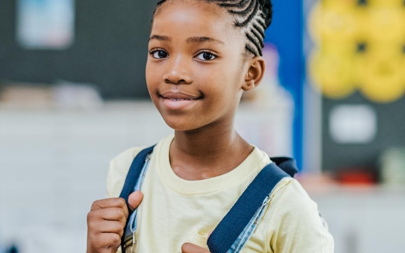 African young girl with blue backpack looking at camera. Pretty and satisfied black schoolgirl with rucksack smiling in class. Portrait of beautiful school girl standing in classroom.