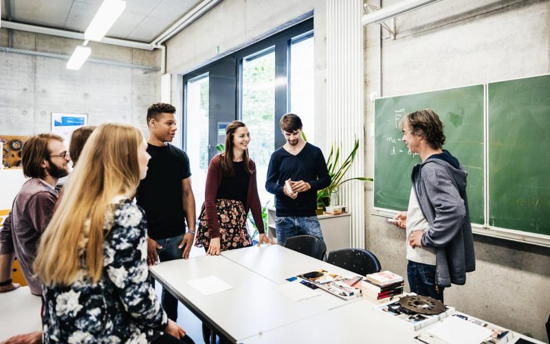 A university tutor talking with a group of students in front of a blackboard during a class.