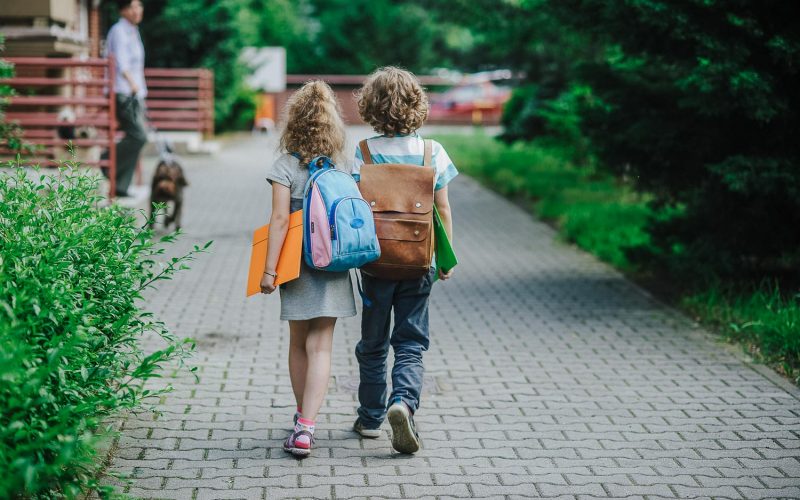 Two pupils of primary school go hand in hand. Boy and girl with school bags behind the back. Beginning of school lessons. Warm day of fall. Back to school.  Little first graders.