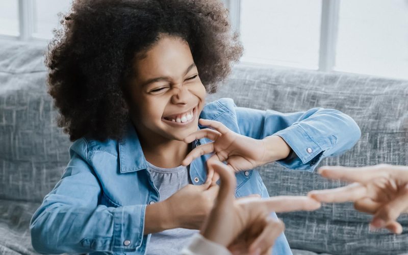 African deaf kid girl and her mother sitting on couch showing symbols with hands using visual-manual gestures enjoy communication at home. Hearing loss disability sign language learning school concept