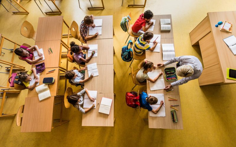 High angle view of large group of elementary students having a class in the classroom. Teacher is assisting one student.