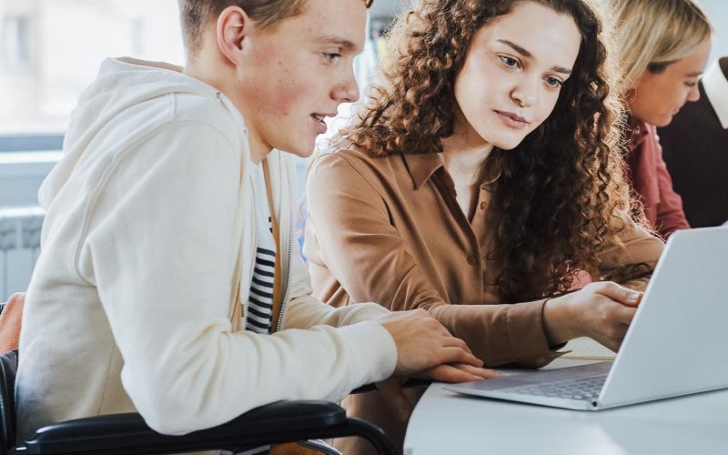 Side view at multi-ethnic group of students using laptop while studying in college, featuring boy using wheelchair