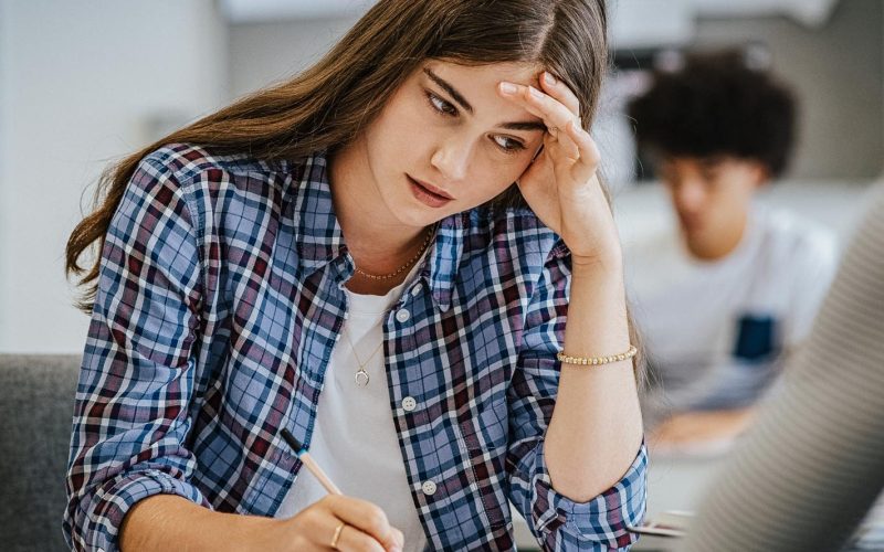 Anxious young woman with hand on head feeling tired while studying at school. College student suffering from headache in classroom. Troubled and stressed girl doing exam that doesnâ€™t know the answers.