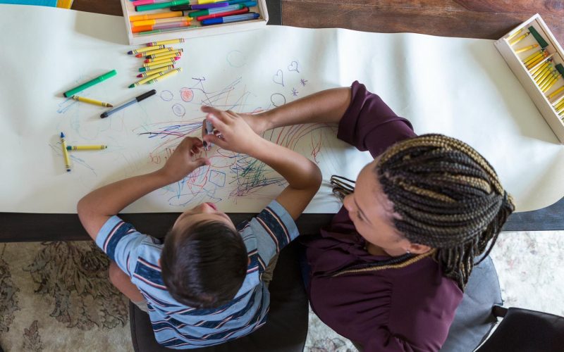 High angle view of elementary age boy with Down Syndrome coloring with female therapist during play therapy session.