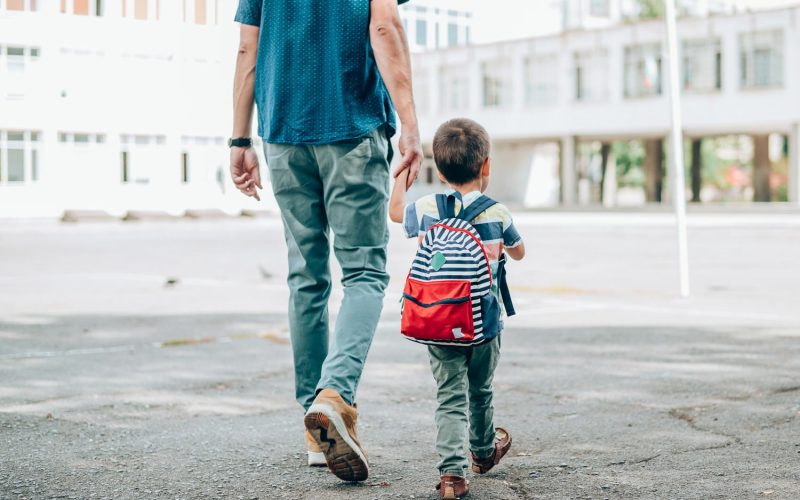 Rear view of father who leads a little boy hand in hand to the school. Father and son with backpack walking in schoolyard.