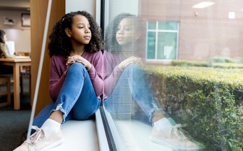 The young girl sits on the windowsill at the library as she looks out the window and daydreams.