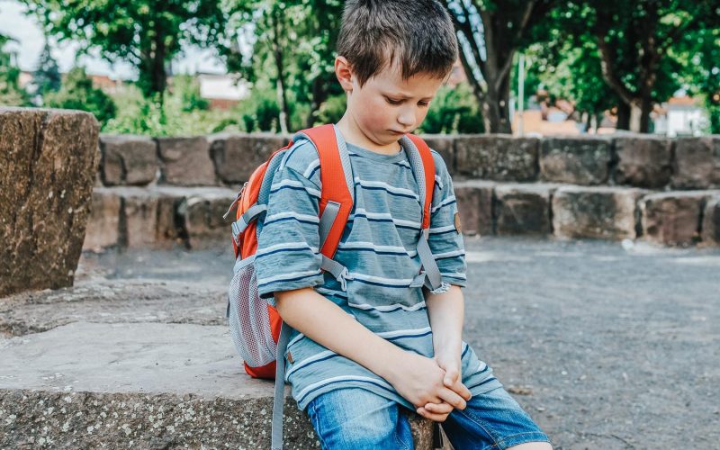 A sad schoolboy sits in the school yard with a backpack on his back. Children's problems at school. The boy does not want to go to school