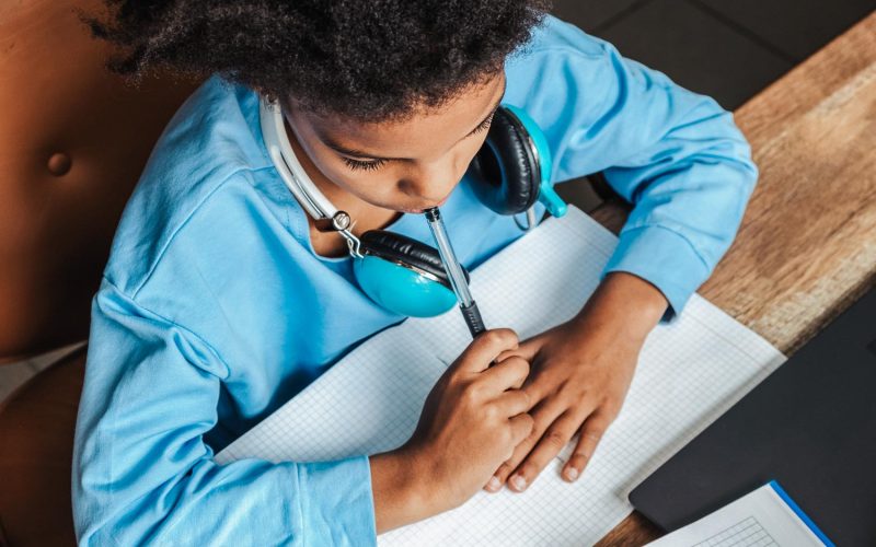 African-American girl doing homework,making notes,using headphones and laptop at home.Back to school concept.School distance education at home,home schooling,e-learning,diverse people.