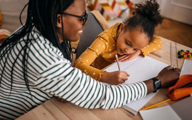 Mid adult mother with dreadlocks assisting daughter in writing homework on table at home