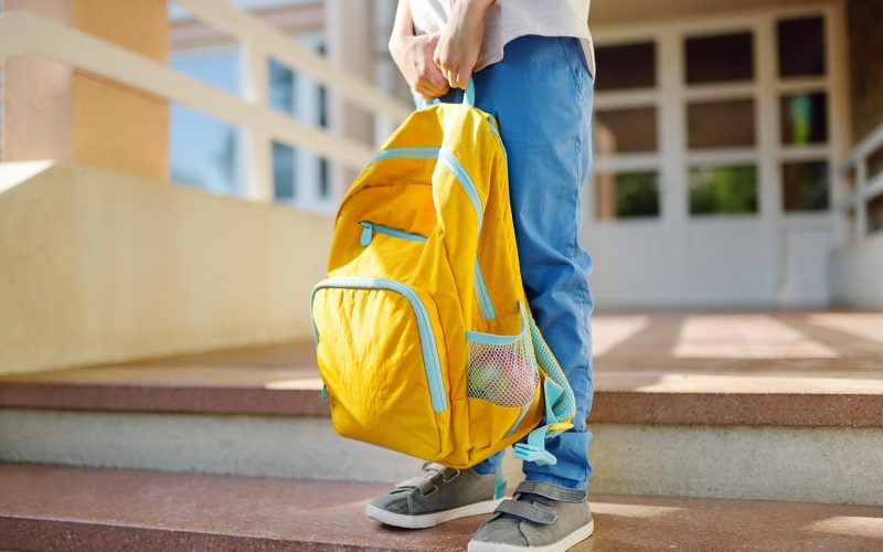 Little student with a backpack on the steps of the stairs of school building. Close-up of child legs, hands and schoolbag of boy standing on staircase of schoolhouse. Kids back to school concept.