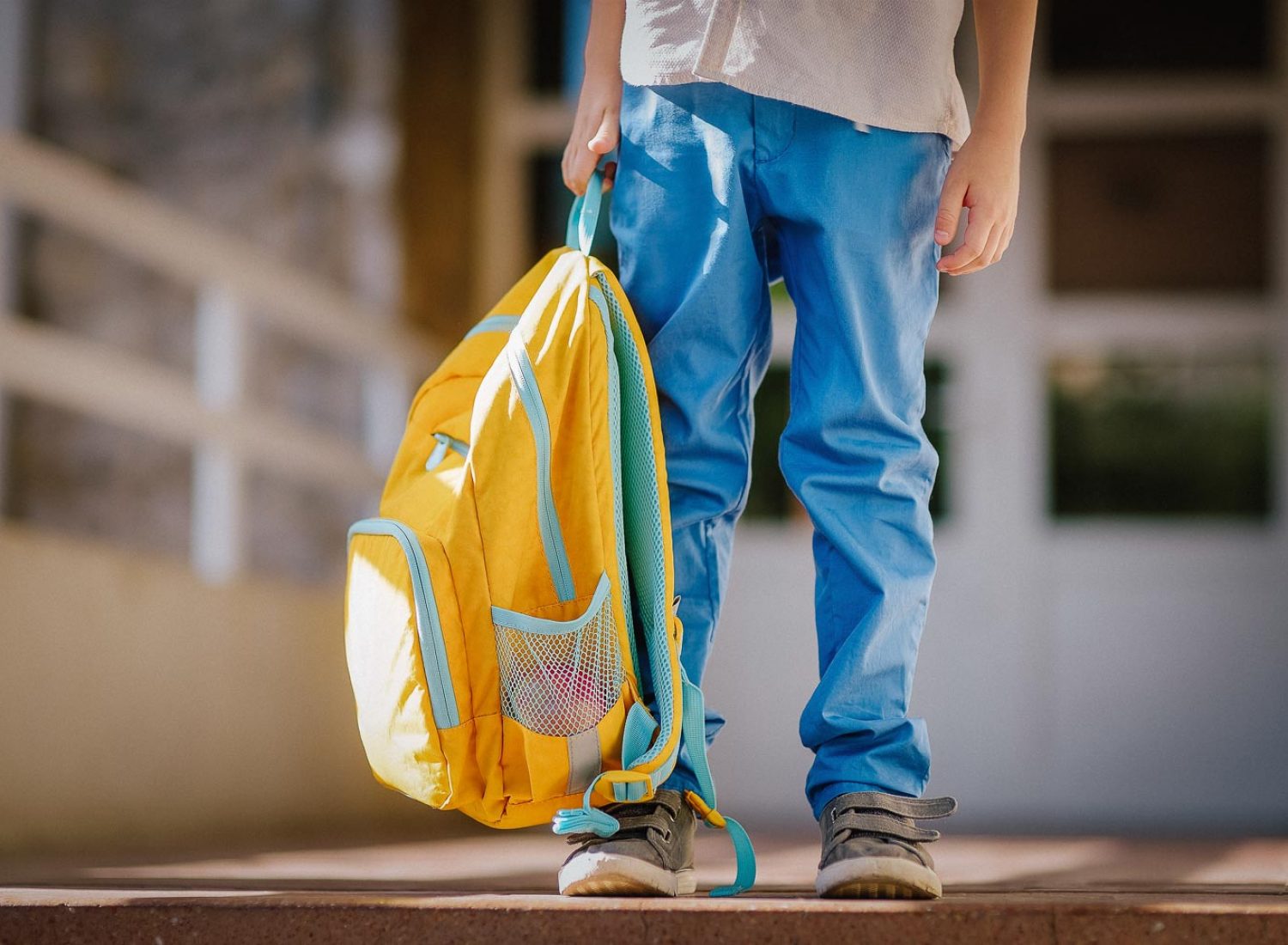 Little student with a backpack on the steps of the stairs of school building. Close-up of child legs, hands and schoolbag of boy standing on staircase of schoolhouse. Kids back to school concept.