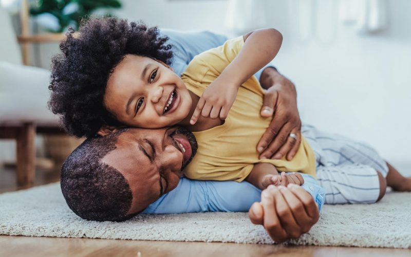 Black father in living room have a fun playing together on warm floor at home.Happy African family dad hugging, embracing his lovely child while lying on the floor.Sweet moments of fatherhood concept.