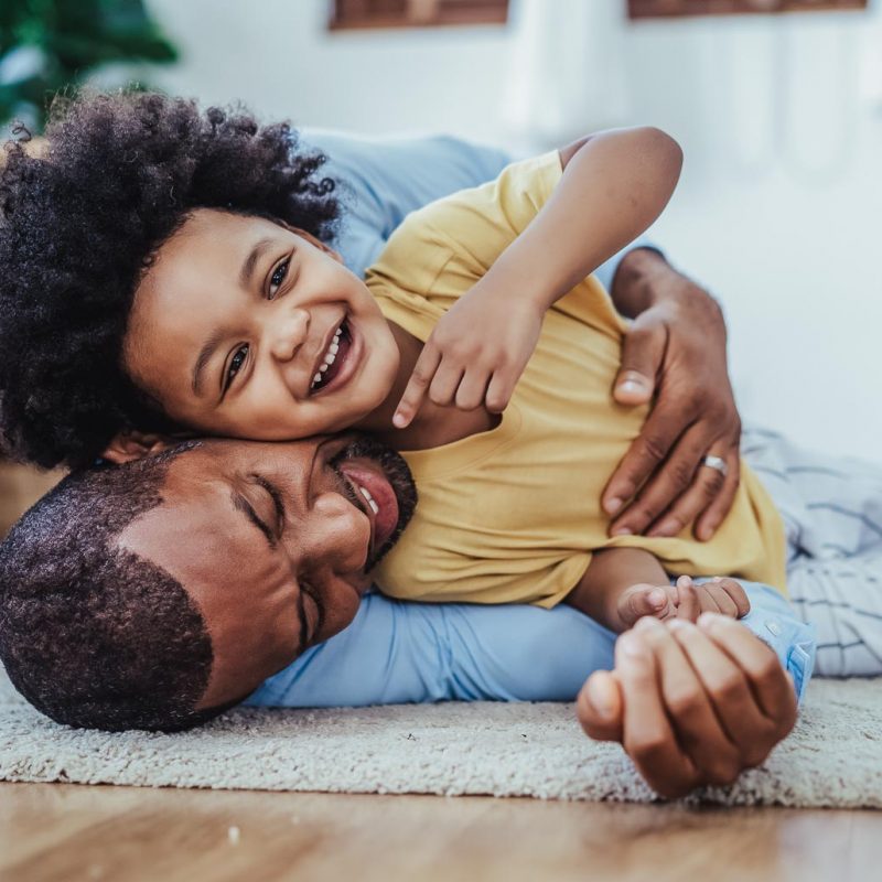 Black father in living room have a fun playing together on warm floor at home.Happy African family dad hugging, embracing his lovely child while lying on the floor.Sweet moments of fatherhood concept.