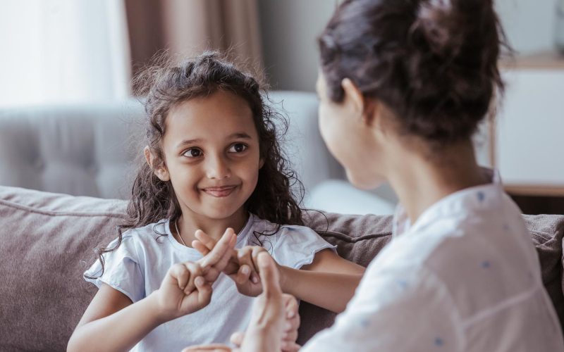 Happy cute Indian kid girl talking to mother, using hands, fingers, speaking sign languages, sitting on sofa at home. Female teacher, therapist teaching child with deafness to communicate