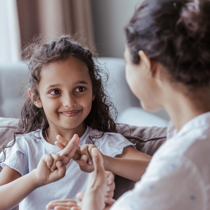 Happy cute Indian kid girl talking to mother, using hands, fingers, speaking sign languages, sitting on sofa at home. Female teacher, therapist teaching child with deafness to communicate