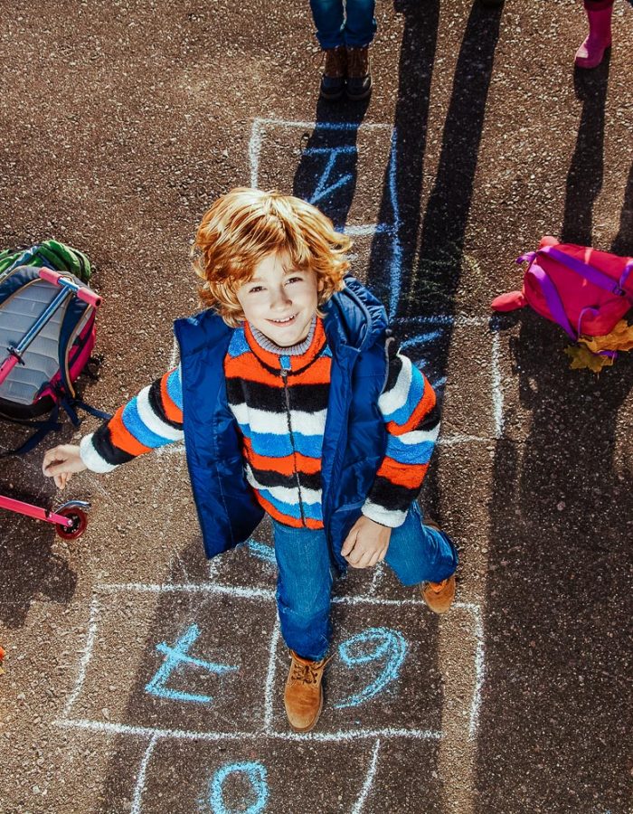 Nice blond boy jumping over hopscotch game after school with bags and scooter laying near