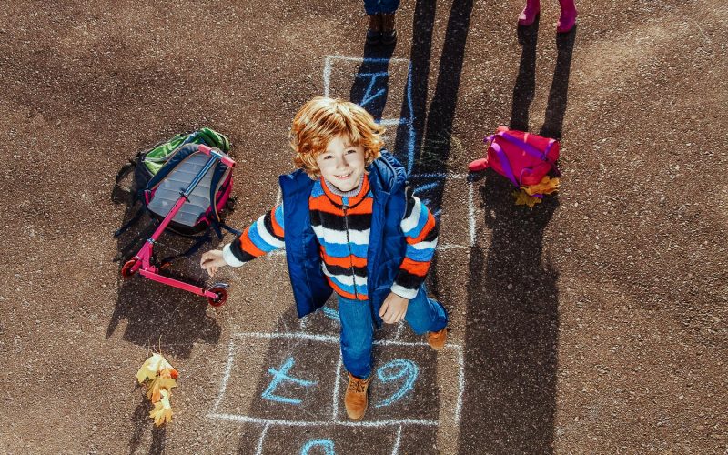 Nice blond boy jumping over hopscotch game after school with bags and scooter laying near