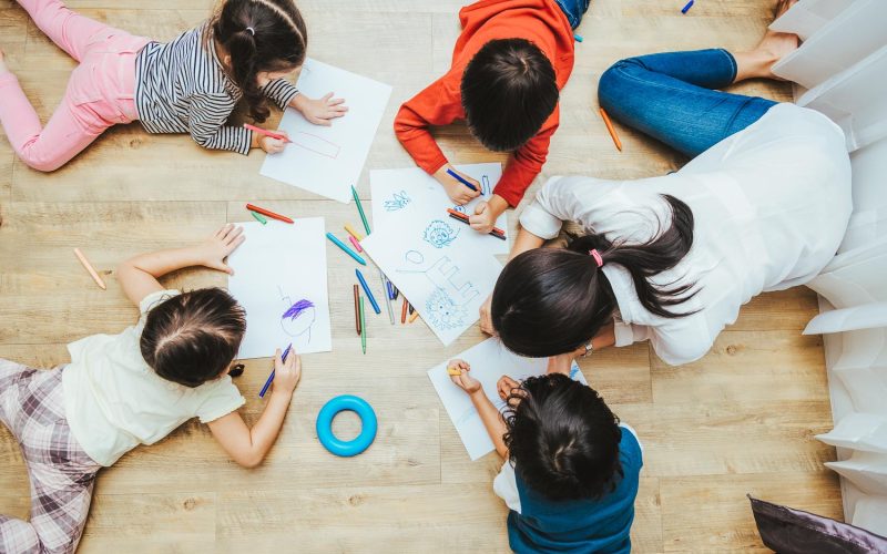 Top view Family happy children group kids boy and girl kindergarten paint drawing on paper with woman teacher education together at interior playroom or nursery, back to school concept