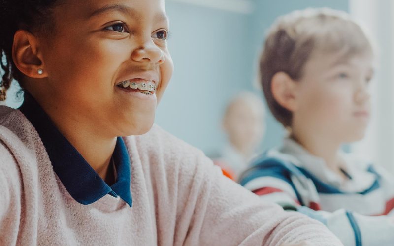In Elementary School Class: Portrait of a Brilliant Black Girl with Braces Smiles, Writes in Exercise Notebook. Junior Classroom with Diverse Group of Children Learning New Stuff