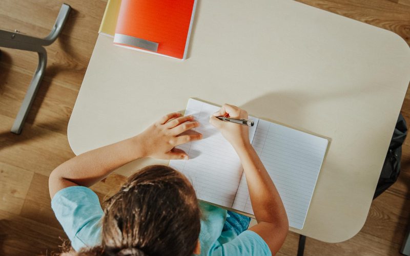 Top View Shot of Elementary School Classroom: Girl Sitting at the School Desk Working on Assignments in Exercise Notebooks.