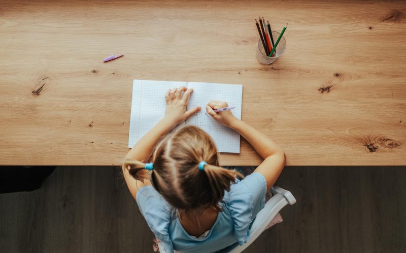 High-angle view of unrecognizable primary little child girl doing homework sitting at home table by window. Smart preschool kid studying alone drawing with pen in bedroom. Homeschooling concept.
