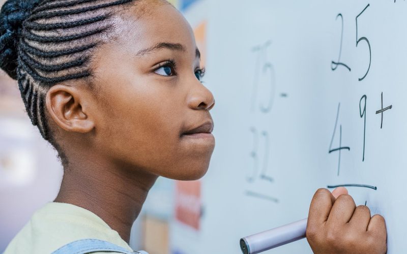 Portrait of african girl writing solution of sums on white board at school. Black schoolgirl solving addition sum on white board with marker pen. School child thinking while doing mathematics problem.