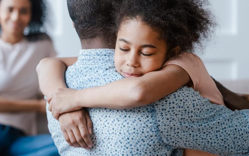 Young african american man holding, embracing, comforting smiling happy calm black cute kid daughter, blurred mother sitting on couch on background, loving supporting family concept.