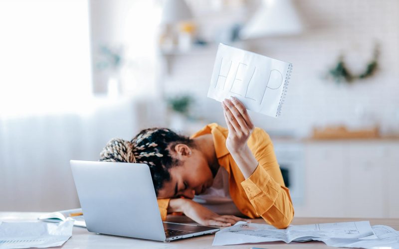 A frustrated African American female student is tired of studying at home, lies at her desk and holds a leaflet with the inscription help. Freelancer young woman need a rest from remotely work