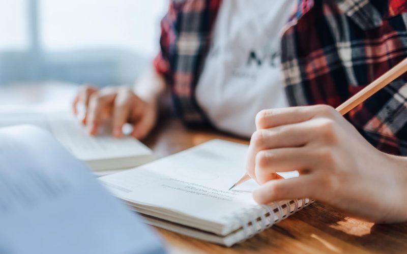 Asian teen girl wearing headphones learning language online, using laptop, looking at screen, doing school tasks at home, writing notes, listening to lecture or music, distance education