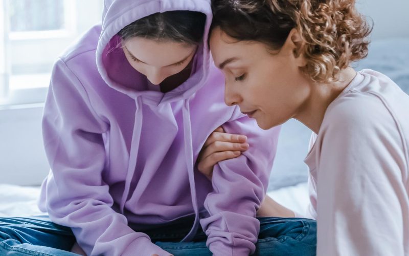Caring mother hugging, comforting depressed upset teenage daughter, stressed unhappy teenager girl wearing hood sitting on bed, thinking about problems, feeling lonely and misunderstood