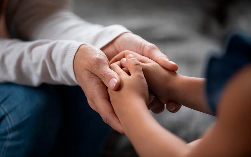 Close up compassionate young foster parent holding hands of little kid girl, giving psychological help, supporting at home. Sincere different generations family sharing secrets or making peace.