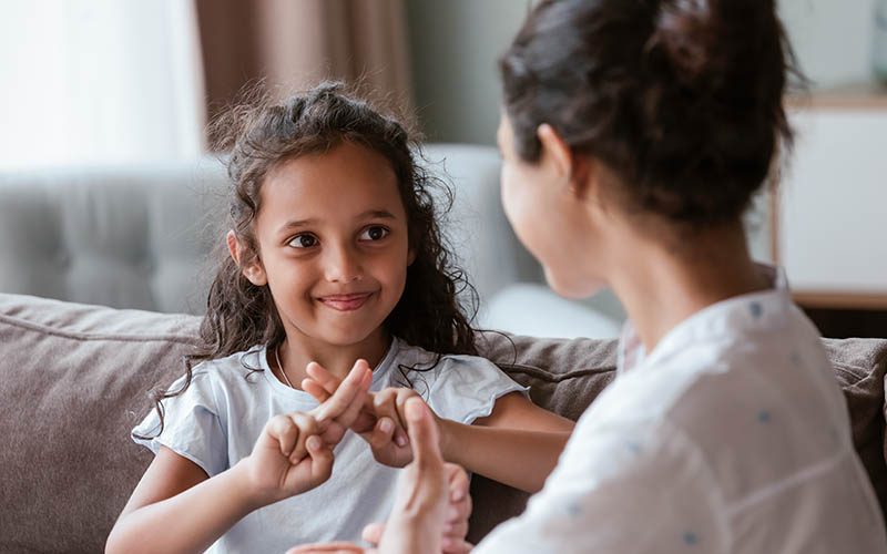 Happy cute Indian kid girl talking to mother, using hands, fingers, speaking sign languages, sitting on sofa at home. Female teacher, therapist teaching child with deafness to communicate