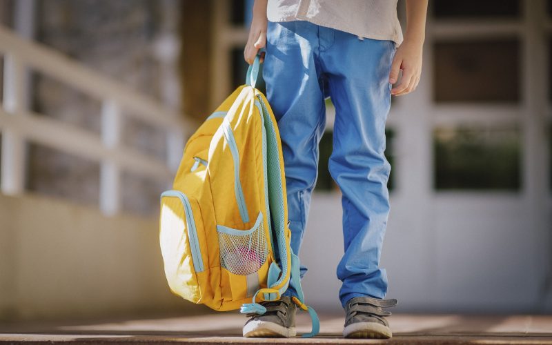 Little student with a backpack on the steps of the stairs of school building. Close-up of child legs, hands and schoolbag of boy standing on staircase of schoolhouse. Kids back to school concept.