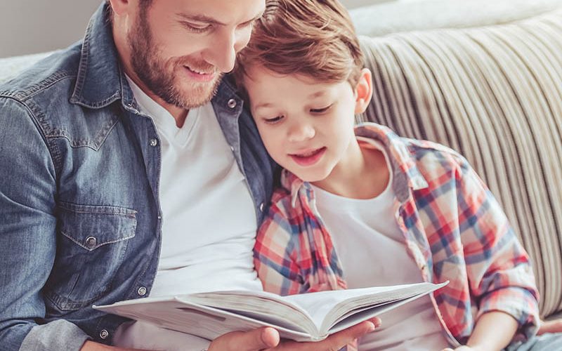 Father and son are reading a book and smiling while spending time together at home
