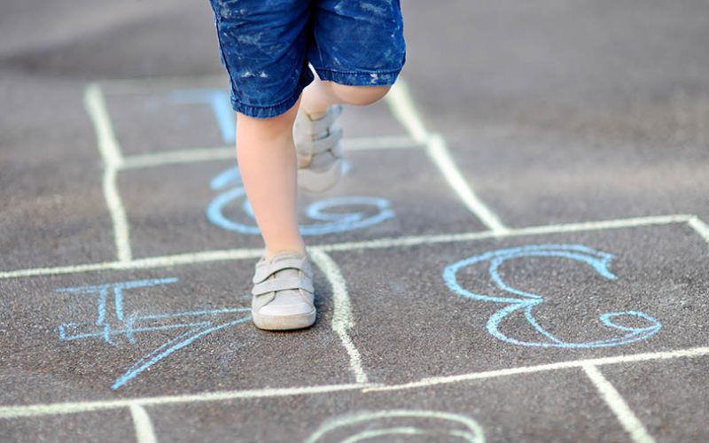 Closeup of little boy's legs and hopscotch drawn on asphalt. Child playing hopscotch game on playground outdoors on a sunny day. Summer activities for children.